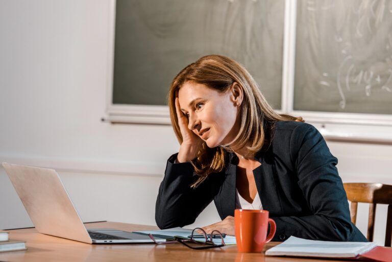 Teacher sitting at a desk with her head in her hands and a glazed look on her face staring at a computer, with glasses and a mug and a binder on the desk beside her with books behind the computer and a blackboard on the wall behind the teacher.